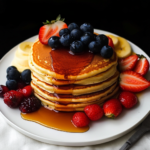 A stack of fluffy pancakes topped with fresh blueberries and strawberries, drizzled with maple syrup, and served with sliced bananas and mixed berries on a white plate.